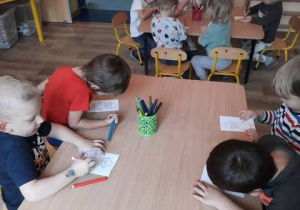 Dzieci siedzą przy stolikach w przedszkolu kolorując Krasnala Hałabałę.
